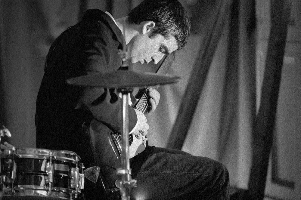 A limited edition Rockarchive print of Noel Gallagher playing guitar beside a drum kit.
