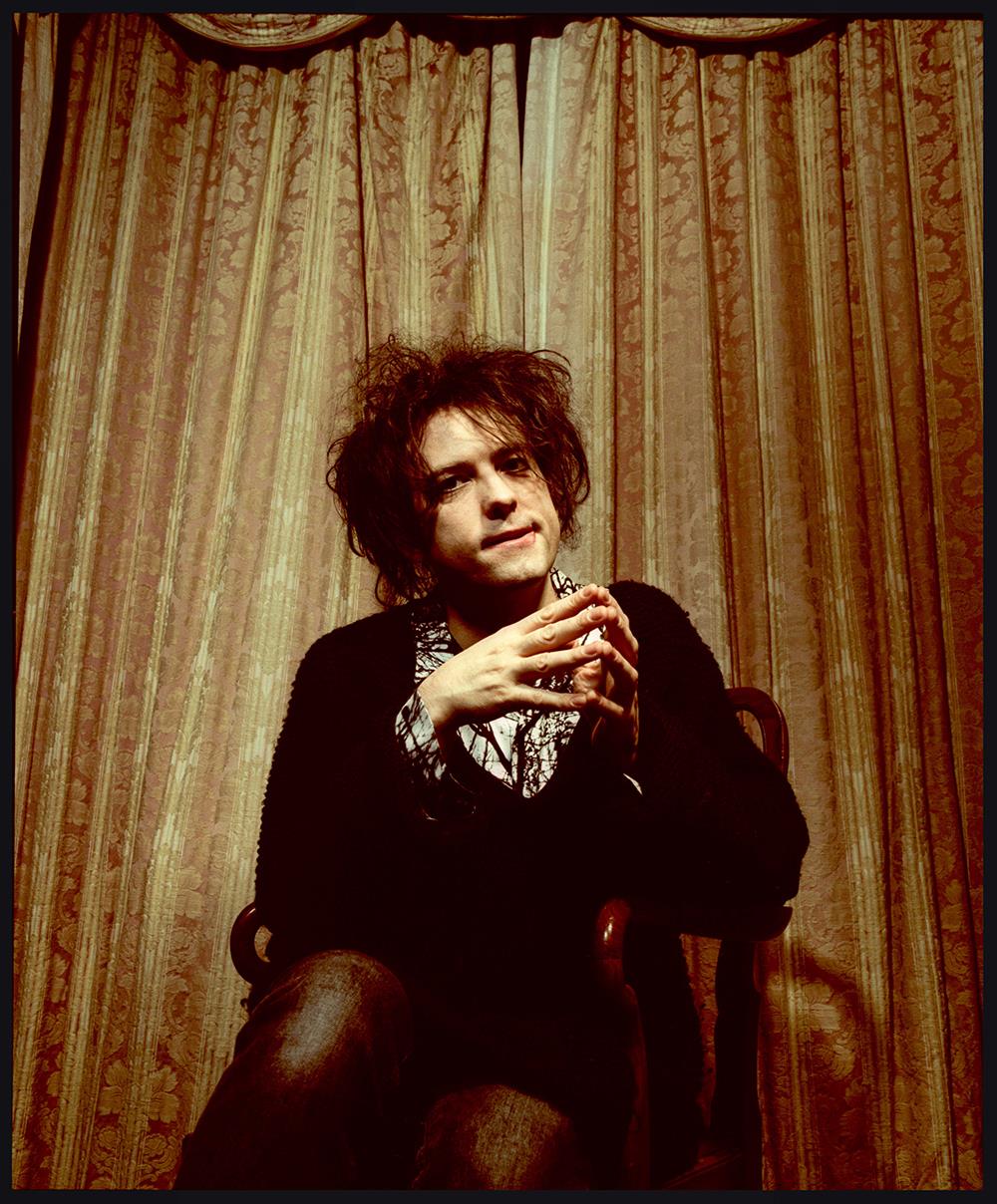 A limited edition colour print of Robert Smith from The Cure sat on a chair in front a pair of curtains.
